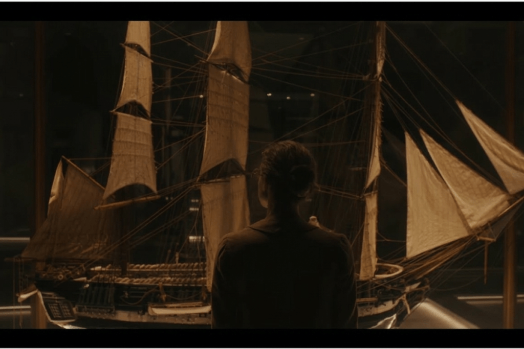 “BTS Swim teaser showing a person with hair tied standing in front of an intricately detailed model sailing ship in dim museum-like lighting.”